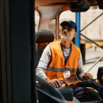 Mature warehouse worker moving pallets with forklift in industrial building.